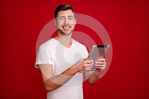 Photo of half turned positive young guy smile hold tablet look camera isolated on dark red color background