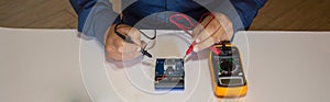 Photo of the hands of a man using a tester to repair an electrical board of a battery pack. Horizontal banner