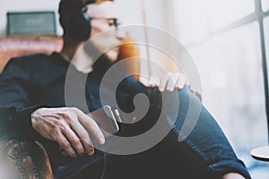 Photo handsome bearded man headphones listening to music modern loft studio.Man sitting in vintage chair looking window,holding sm