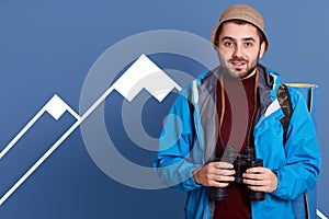 Photo of handsome male traveler spending free time actively, carries binoculars for exploring surroundings, poses over blue studio