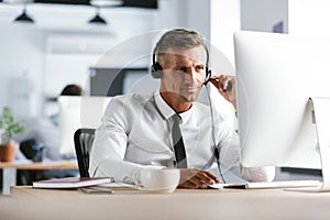 Photo of handsome man 30s wearing office clothes and headphones, sitting by computer in call center