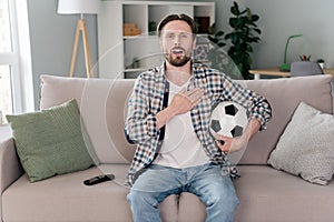 Photo of handsome pretty young man wear plaid shirt sitting sofa watching tv-set arm chest singing anthem indoors house