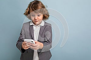 Photo of Handsome surprised boy with curly hair wearing grey suit holding and using phone isolated over blue background