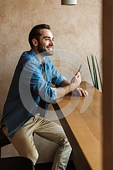 Photo of handsome unshaved man smiling and using cellphone while sitting on chair in cafe indoors