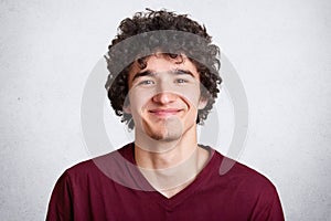 Photo of handsome unshaven guy looks with pleasent expression directly at camera, wears fashionable maroon t shirt, stands against photo