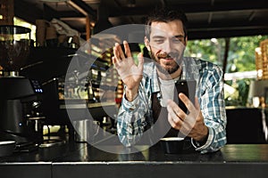 Photo of happy barista guy smiling while using smartphone in cafe or coffeehouse outdoor