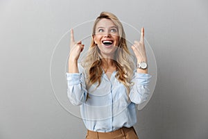 Photo of happy blond businesswoman with long curly hair smiling and pointing fingers upward at copyspace