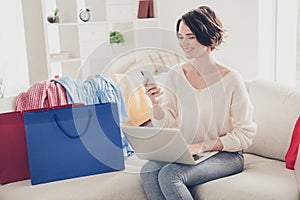 Photo of happy brunette woman sit sofa look phone hold laptop shop online bags indoors relax home living-room