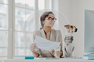 Photo of happy businesswoman works from home on self isolation, holds papers, checks information on computer, wears glasses,