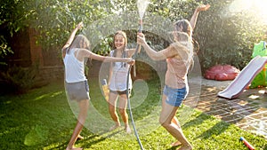 Photo of happy cheerful girls in wet clothes dancing and jumping under water garden hose. Family playing and having fun