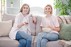 Photo of happy grey haired old lady and blonde woman sit sofa watch tv eat popcorn indoors inside house home