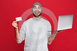 Photo of happy guy holding credit card and silver laptop isolated over red background