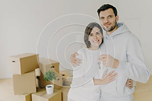 Photo of happy husband and wife buy real estate, cuddle and hold keys, stand in living room with boxes in new home, dressed in