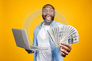 Photo of happy young afro american handsome man posing isolated over yellow wall background using laptop computer holding money