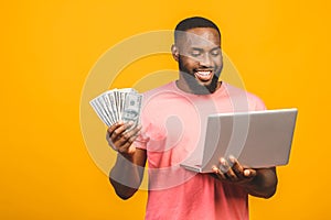 Photo of a happy young afro american handsome man posing isolated over yellow wall background using laptop computer holding money