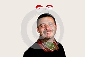 Photo of a happy young man in a black sweater and a red-green scarf in Christmas horns with the face of Santa Claus on his head.