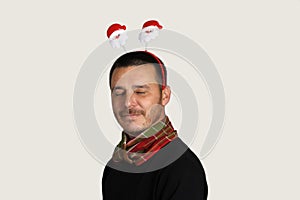 Photo of a happy young man in a black sweater and a red-green scarf in Christmas horns with the face of Santa Claus on his head.