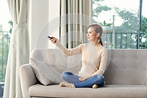 Photo of happy young woman sitting on sofa at home. Looking camera holding remote control watch TV