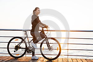 Photo of healthy woman riding bicycle on boardwalk, during sunrise over sea