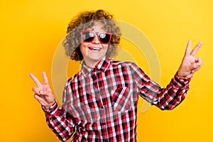 Photo of hipster orange hairstyle teenager boy show v-sign wear eyewear red checkered shirt isolated on yellow color