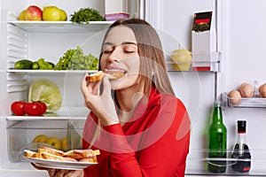 Photo of hungry young cute woman eats with appetite tasty sausage sandwhich, comes after work, stands near opened refrigerator, cl