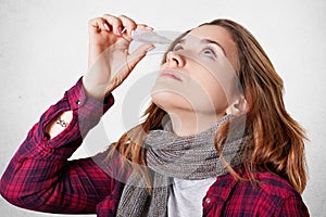 Photo of ill female has bad eyesights, uses spray for dripping eyes, wears knitted scarf, over white background. Sick wom