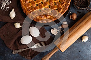 The photo illustrates homemade pie with mushrooms isolated on dark background. The brown napkin, two eggs. garlic, fork, rolling