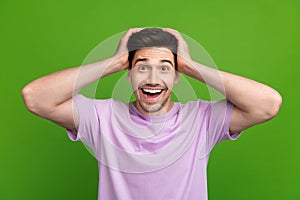 Photo of impressed ecstatic man with brunet hair dressed purple t-shirt hold arms on head staring isolated on green
