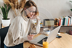 Photo of joyful blonde woman holding credit card and using laptop