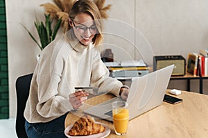 Photo of joyful blonde woman holding credit card and using laptop