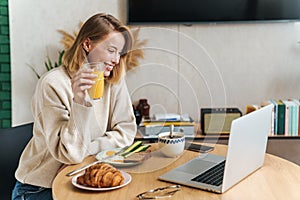 Photo of joyful blonde woman using laptop while having breakfast