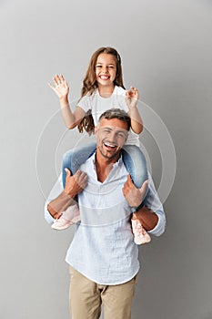 Photo of joyful family smiling at camera while little girl having fun and sitting on the neck of her happy father, isolated over