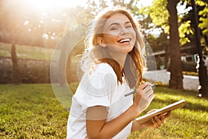 Photo of joyful woman 20s with broad smile sitting on green grass in park with legs crossed during summer day, and writing down n