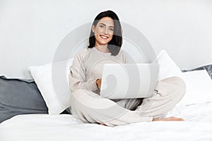 Photo of joyful woman 30s using laptop, while sitting in bed with white linen in cozy apartment