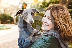 Photo of joyful woman with dog, feels fun, carries her lovely Italian Greyhound puppy, expresses affection, have good relationship