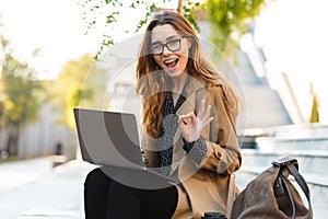 Photo of joyful woman working on laptop while sitting on bench in city street
