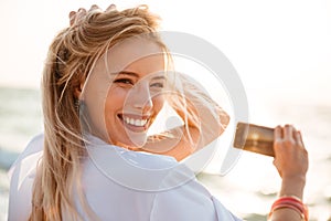 Photo of joyous blonde woman 20s smiling, and taking photo of sunrise above sea, while walking on summer beach