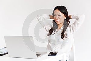 Photo of joyous chinese businesswoman with long dark hair sitting in office and working with documents and laptop, isolated over