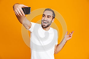 Photo of joyous guy 30s in casual wear laughing and taking selfie on cell phone, isolated over yellow background