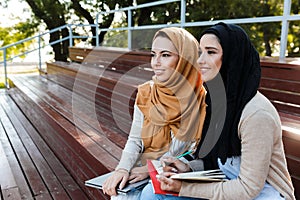 Photo of joyous islamic girls wearing headscarfs sitting on bench in park