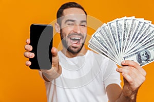 Photo of joyous man 30s in casual wear holding cell phone and fan of money, isolated over yellow background