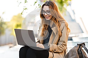 Photo of joyous woman working on laptop while sitting on bench in city street