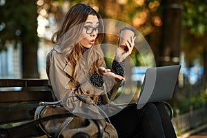 Photo of joyous woman working on laptop while sitting on bench in sunlit alley