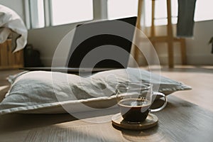 Photo of laptop showing black empty screen putting on white pillow, on wooden floor in cozy room with black coffee cup.