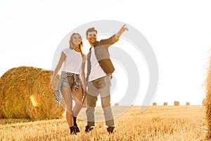Photo of lovely couple man and woman walking through golden field with bunch of haystacks, during sunny day