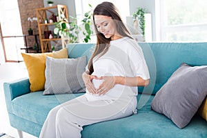 Photo of lovely dreamy cute gentle mama pregnant girl dressed white pajama sitting on sofa touching belly waiting baby