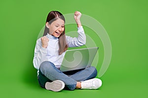 Photo of lucky pretty schoolgirl wear blue turtleneck sitting floor looking modern device rising fists isolated green