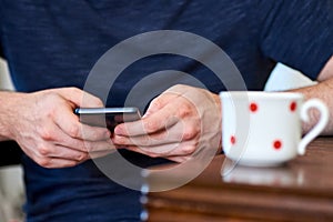 Photo of a man`s hand holding a cell phone. He is sitting in the interior of the apartment and writing a text message on the