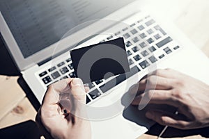 Photo Man Showing Blank Black Business Card and Using Hand Modern Laptop on Wood table Blurred Background. Mockup Ready