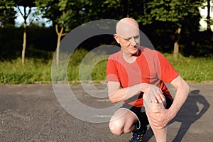 Photo of mature, elderly man sitting in the park and touching his knee by the pain during the day.